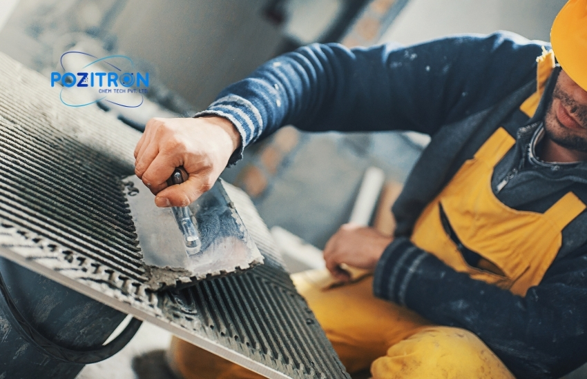 A construction worker in yellow overalls applies adhesive to a tile using a notched trowel, with Pozitron Chem Tech Pvt. Ltd. logo in the top left corner.